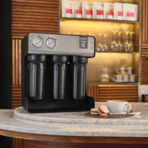 Commercial espresso machine with three filter holders and gauges on a marble counter, beside a cup of coffee and macarons in a cafe setting.